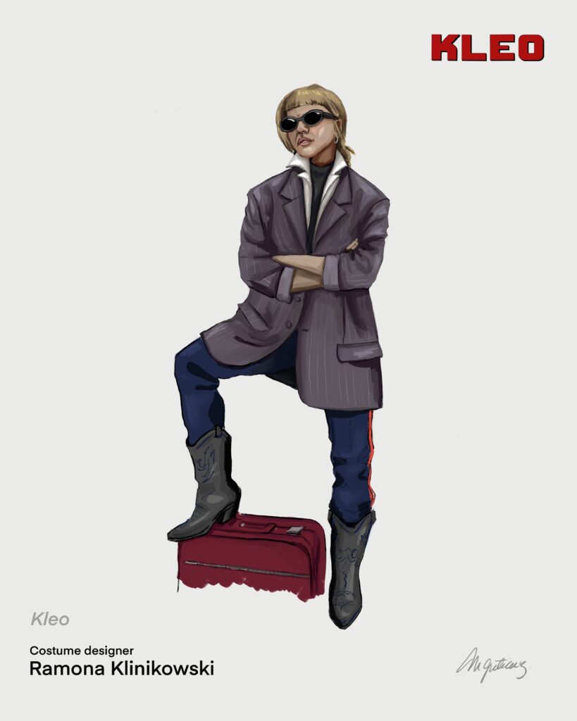 Costume illustration of Kleo: a young woman in sunglasses, a grey oversized jacket, navy trousers with a red stripe and cowboy boots, one foot on a red suitcase